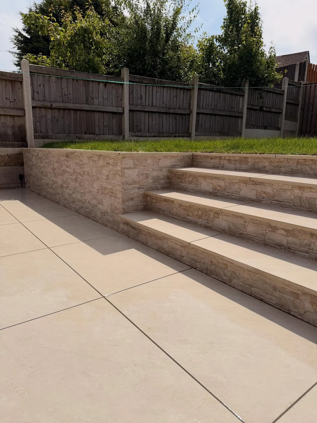 Porcelain patio with broad steps and a stone-effect retaining wall leading to the raised lawn